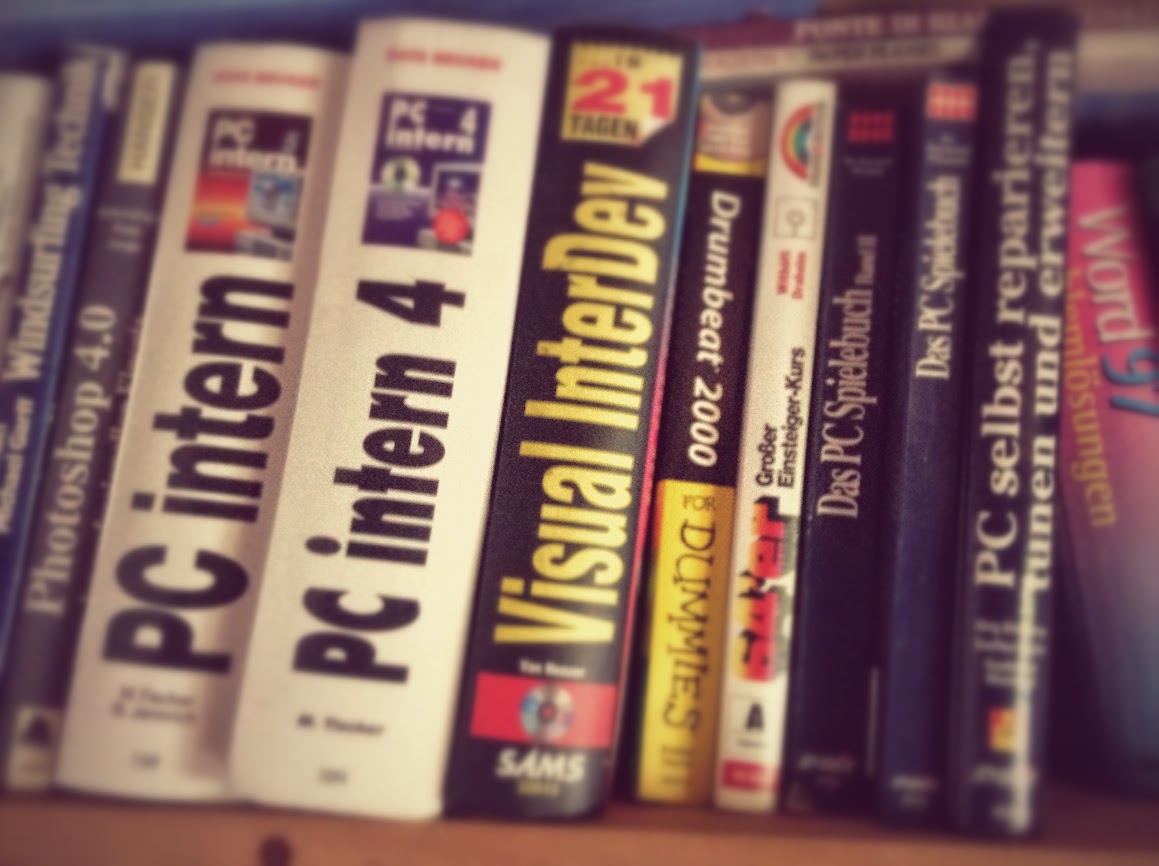 A row of vintage computer programming books from the 1990s and early 2000s on a bookshelf, including titles about PC internals and Visual InterDev. The spines are visible in slightly blurred focus, showing white, black and yellow text on various colored backgrounds.